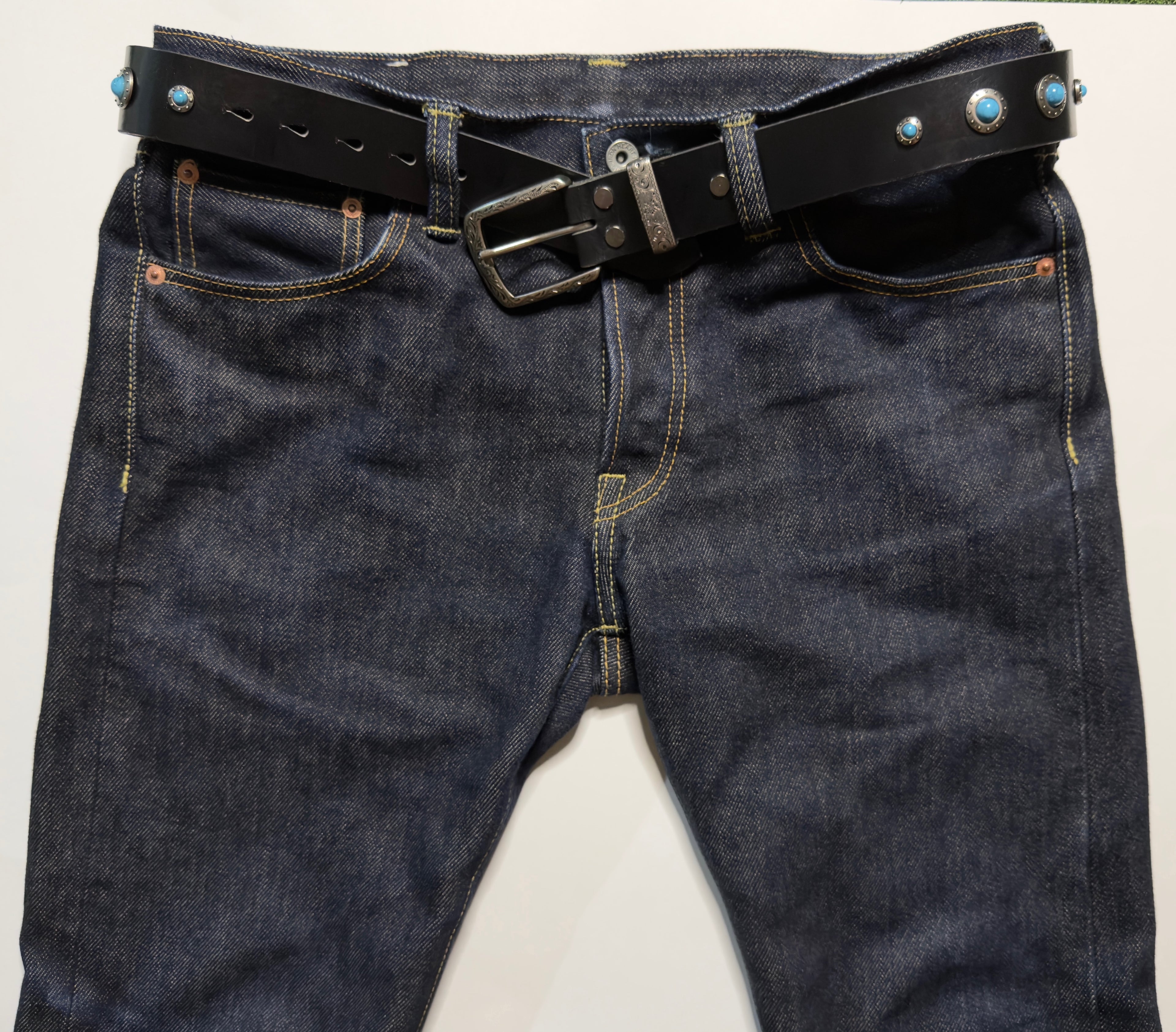 Dark blue jeans with a black belt studded with turquoise conchos on a white background