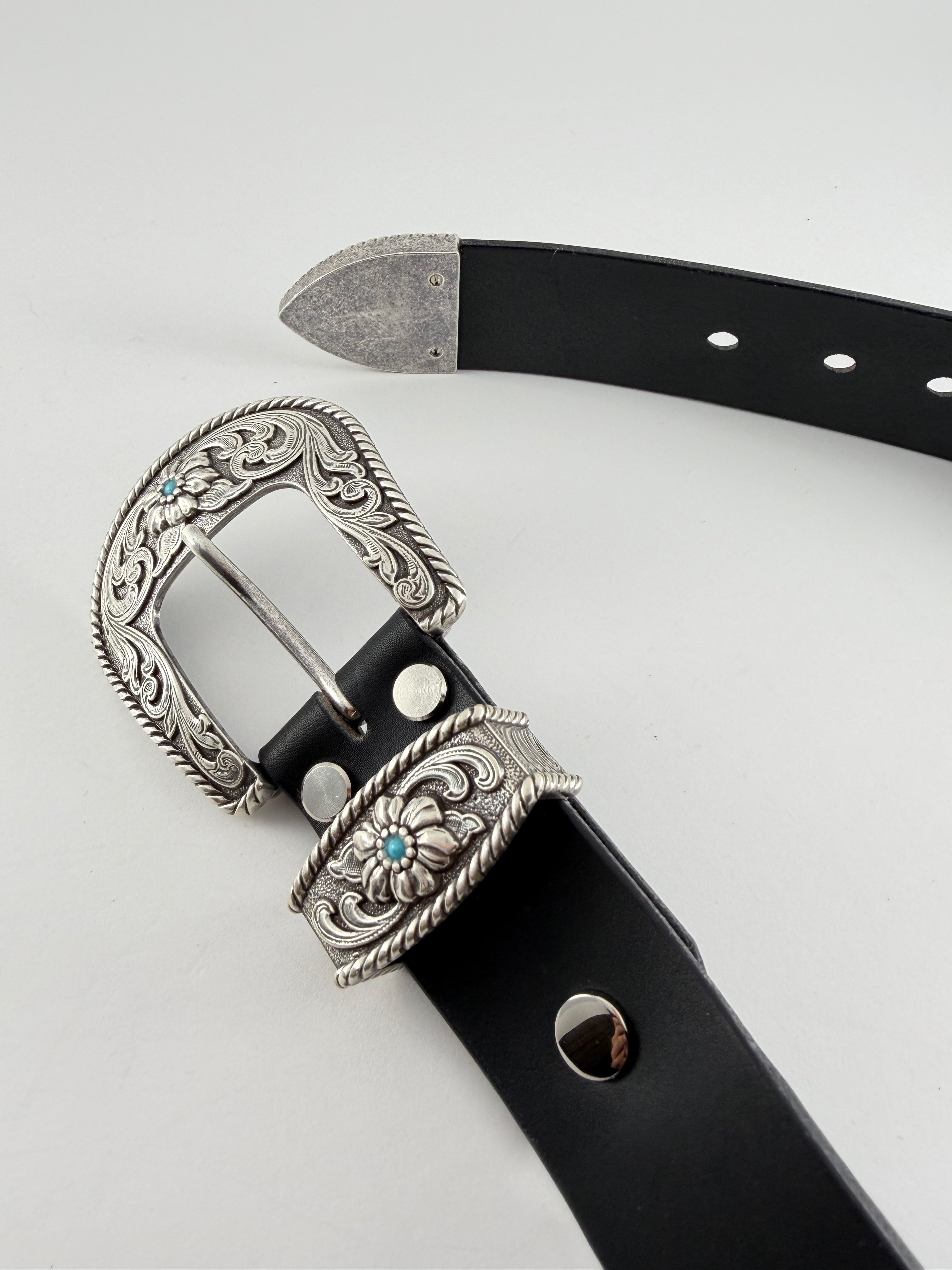 Black western cowboy belt with silver buckle and keeper adorned with turquoise stones on a light gray background