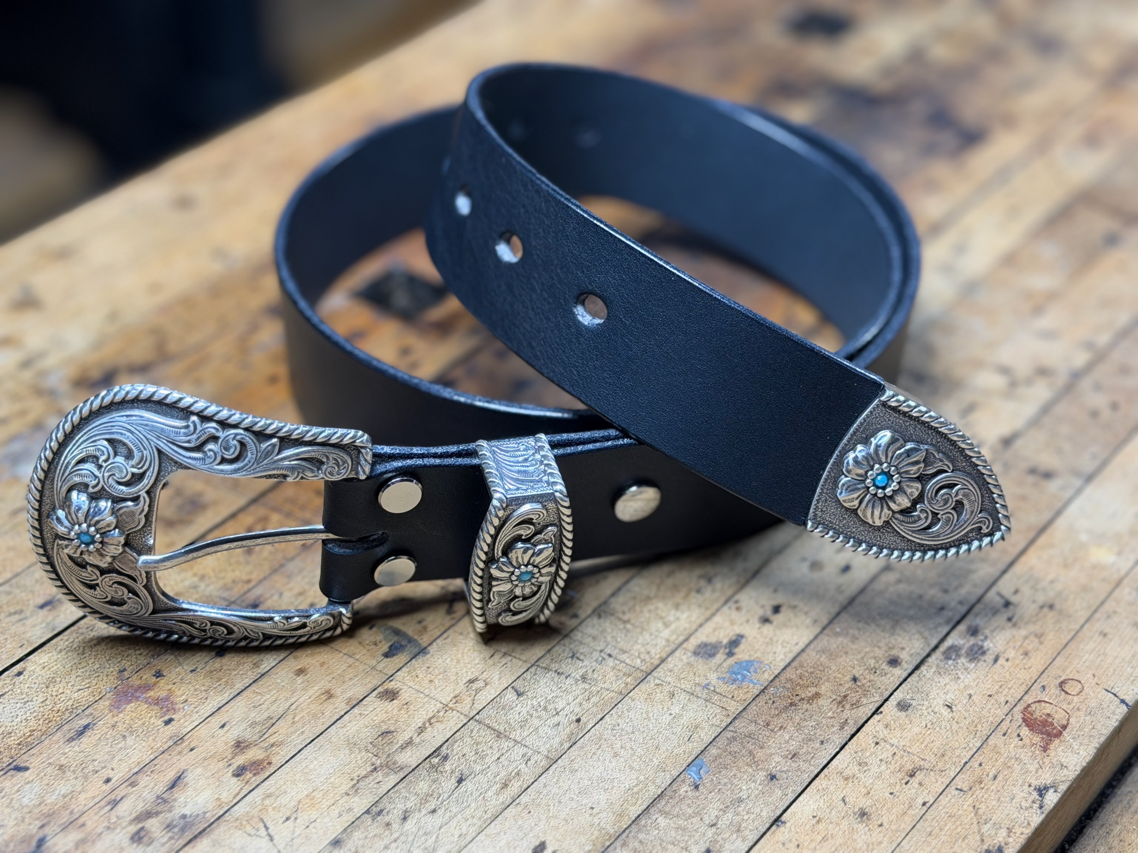 Black western cowboy leather belt with silver buckle and keeper adorned with turquoise stones on a wooden workbench