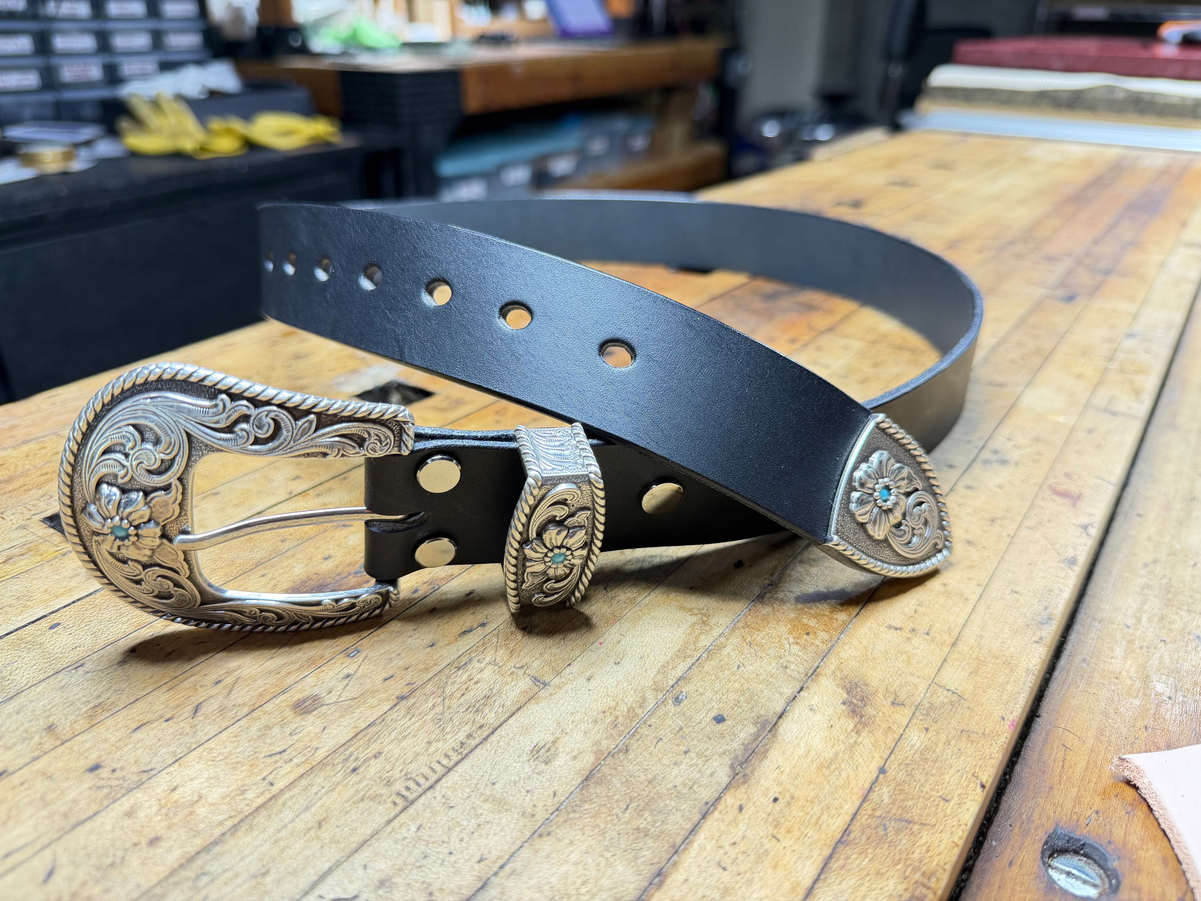Black western cowboy leather belt with silver buckle and keeper adorned with turquoise stones on a wooden workbench