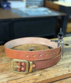 Brown leather belt with brass buckle, keeper and copper rivets on a wooden workbench.