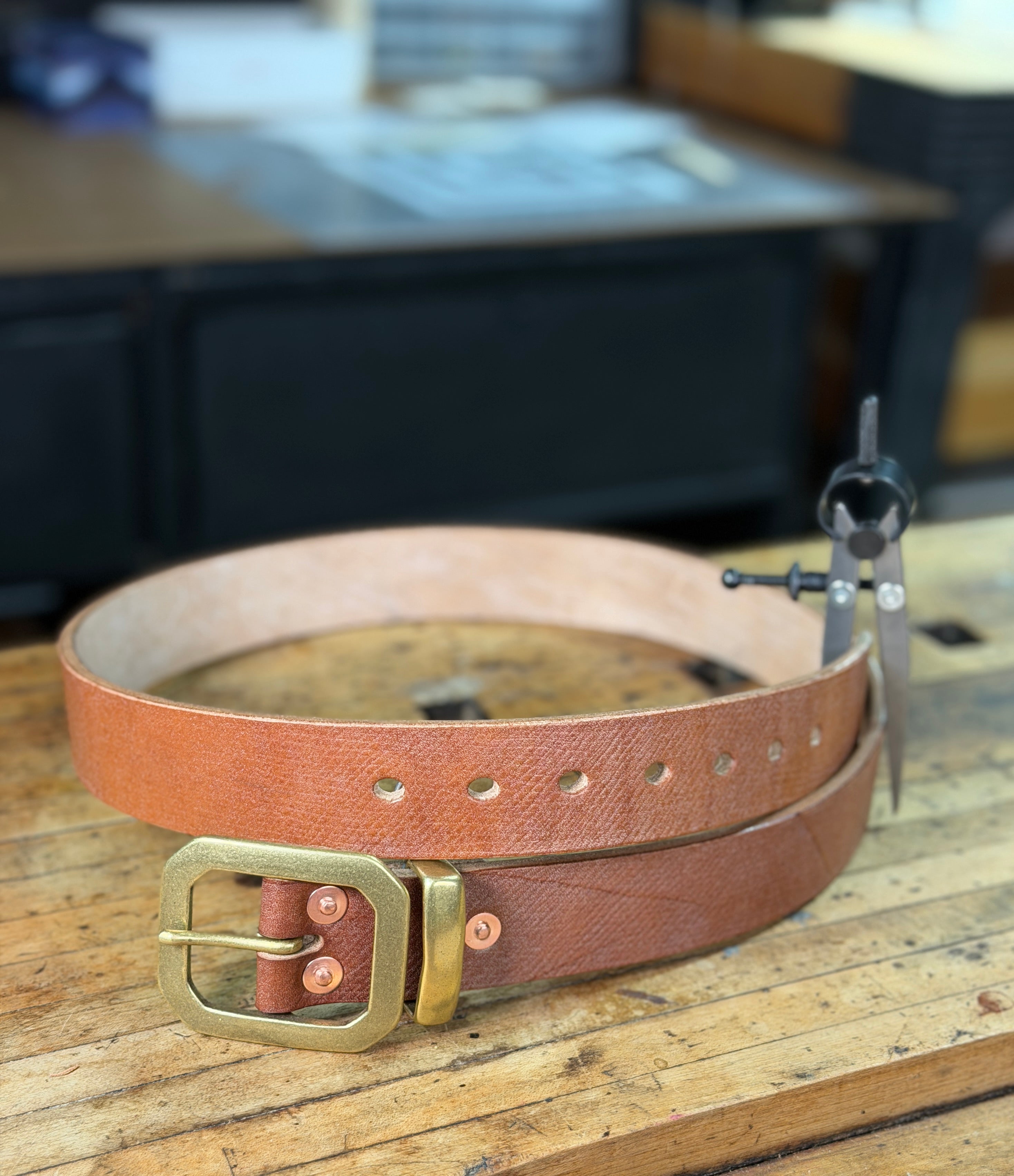 Brown leather belt with brass buckle, keeper and copper rivets on a wooden workbench.