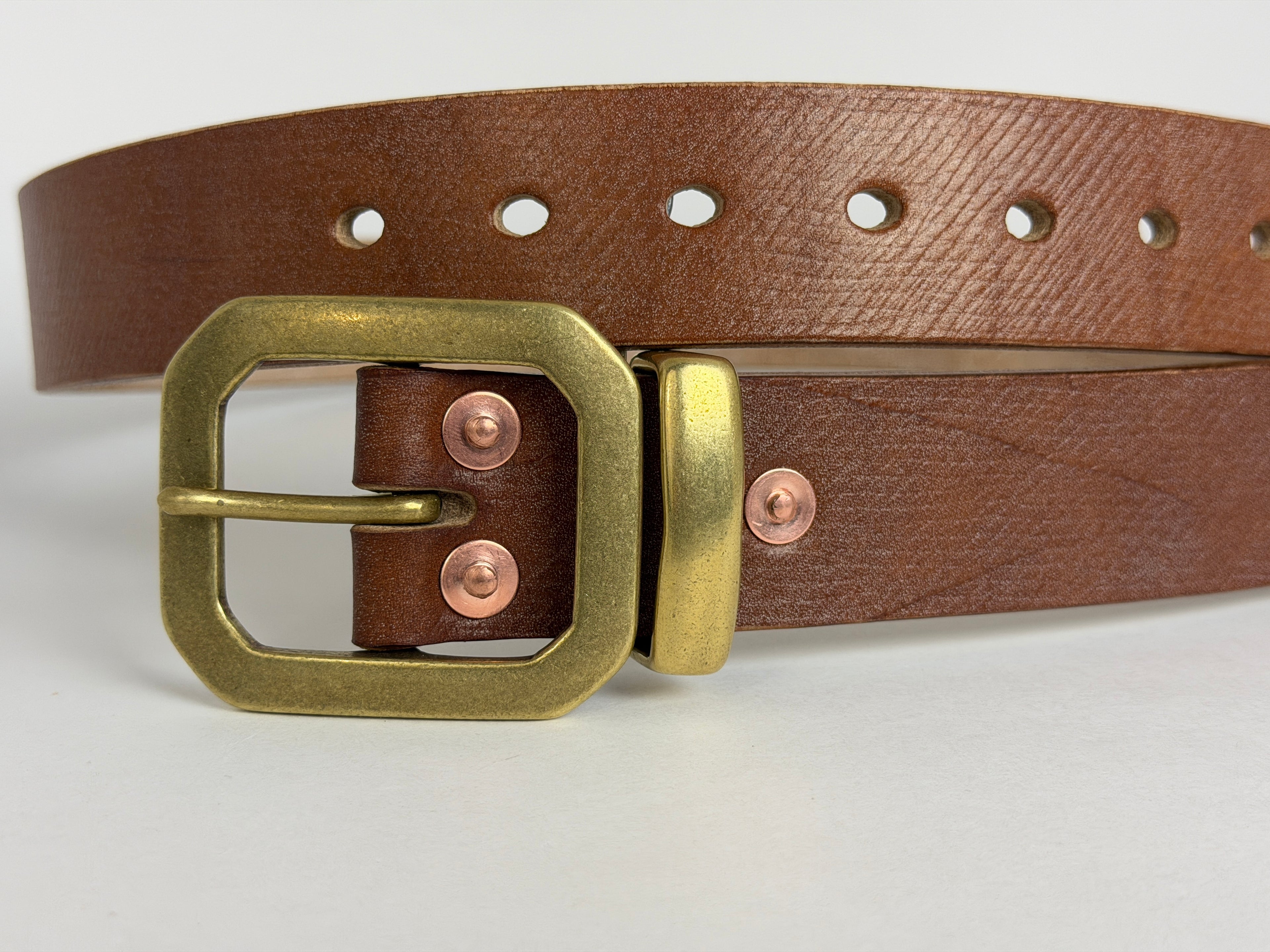 Closeup from view of a brown leather belt with a brass buckle, keeper and copper rivets on a white background.  The 7 punched holes are also in focus.