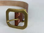 Closeup frontal view of an octagonal solid brass buckle on a brown leather belt on a white background.