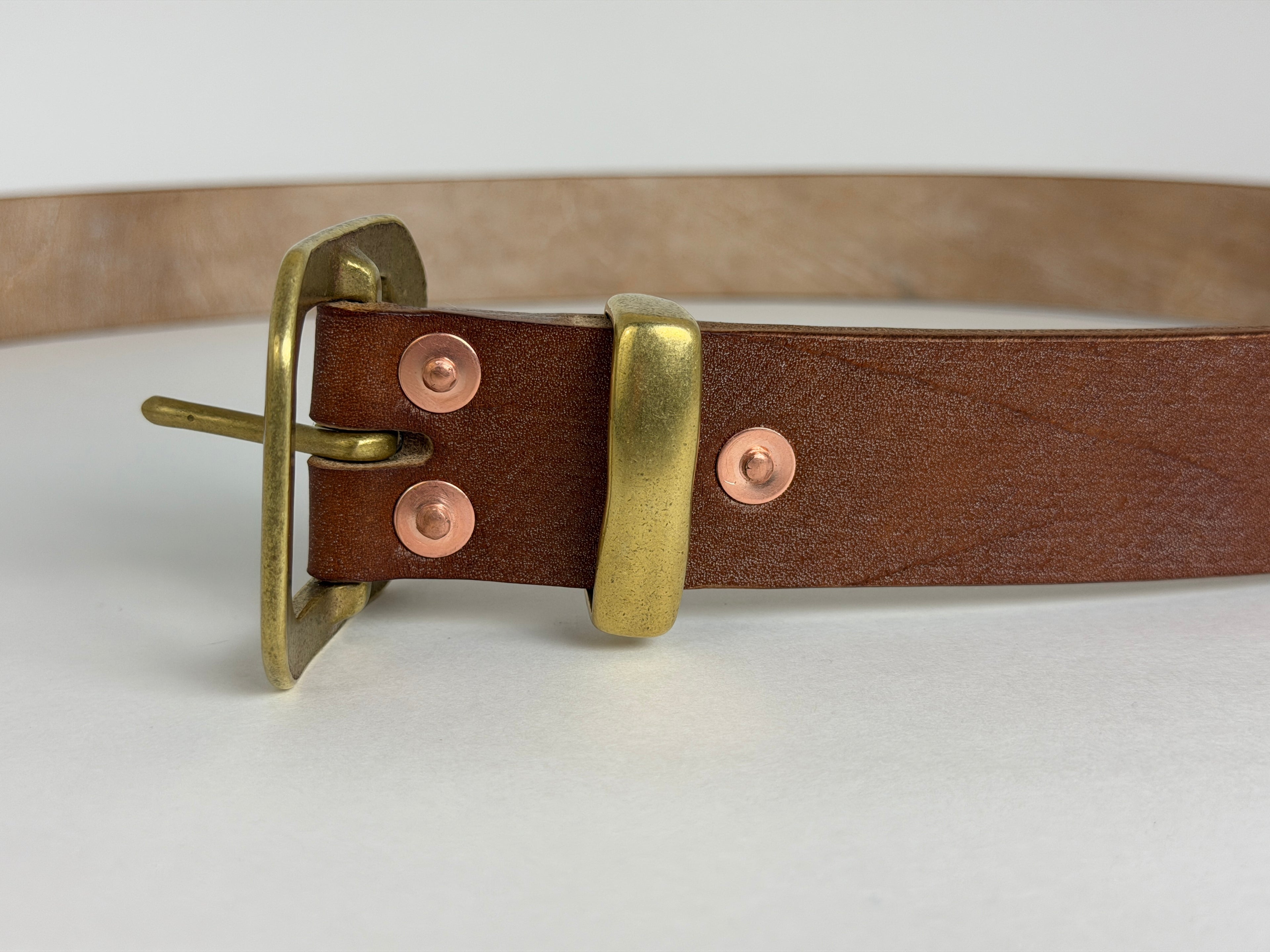 Closeup front view of a brown leather belt with a brass buckle, keeper and copper rivets on a white background.