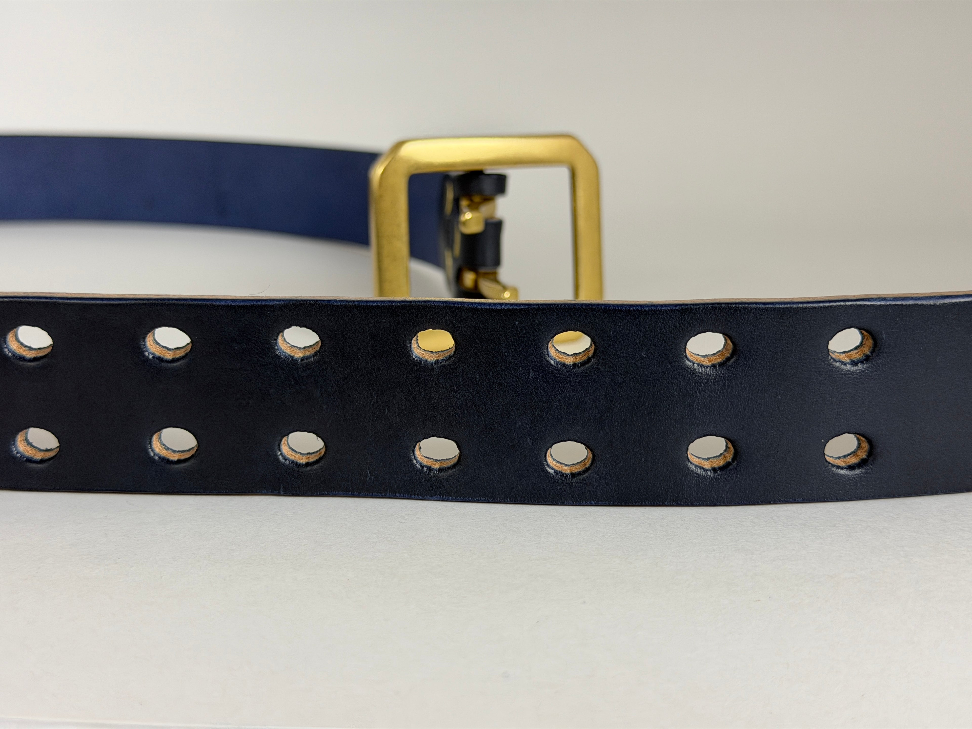 Close-up of a navy blue leather belt with double-pronged holes and a brass buckle on a white background