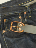 Close-up of a dark indigo denim waistband with contrast yellow stitching, a dark green leather belt with red copper horseshoe-shaped buckle and copper rivets.