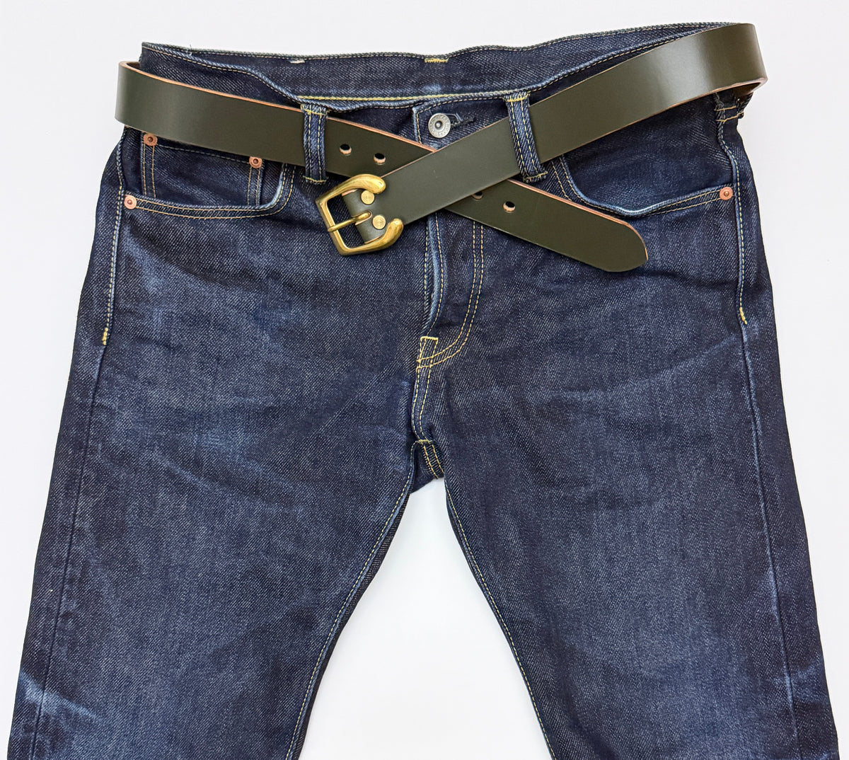 Dark green leather belt with a brass horseshoe  buckle, styled crossed over the waist of dark blue denim jeans on a white background.
