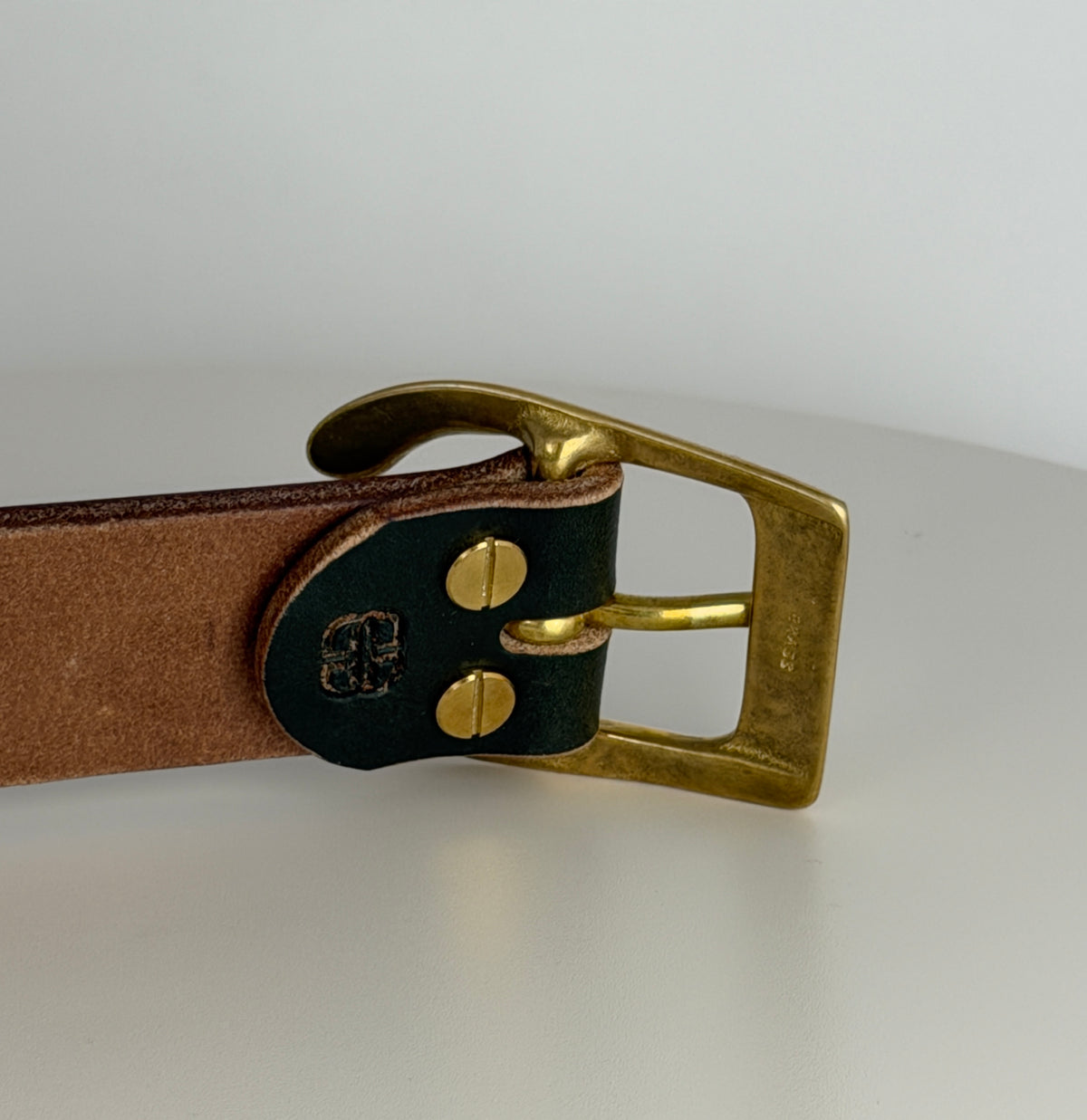 A close-up of the back of a dark green belt on a white background.  This picture highlights how the brass buckle is attached using brass Chicago screws.