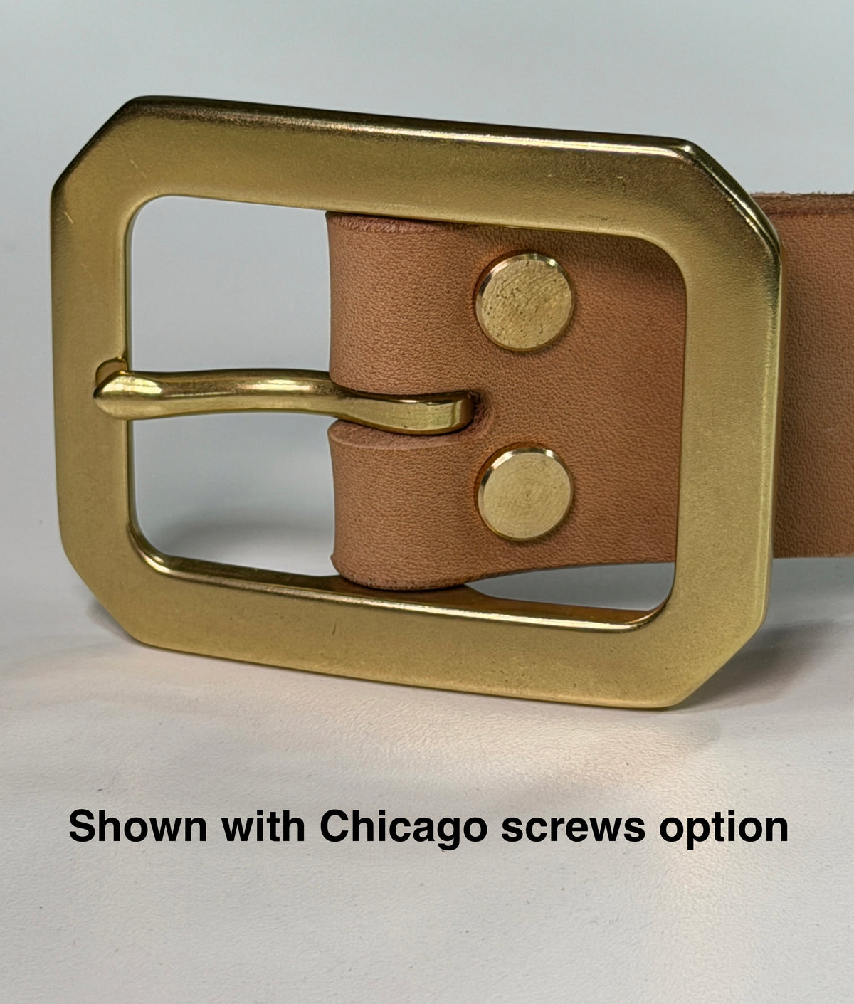 A close-up of a natural leather belt with a large Garrison brass buckle attached with Chicago screws, shown on a light gray background.  There is a label at the bottom of the picture with the words 'Shown with Chicago screws option'.