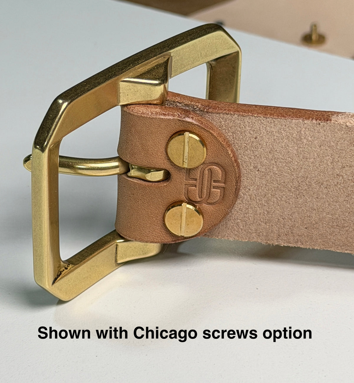 Close-up of a natural leather belt with a solid brass buckle attached with two brass Chicago screws on a white workbench.  There is a caption with the words 'Shown with Chicago screws option' at the bottom of the picture.
