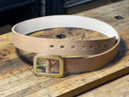 A natural leather belt with a thick brass buckle and red copper rivets, coiled on a rustic wooden workbench.