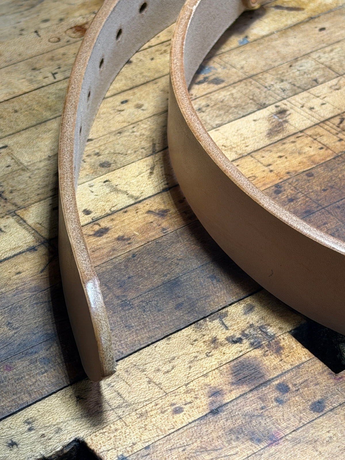 A close-up of the edges of a natural leather belt on a rustic wooden workbench.  The picture highlights the smooth waxed and burnished edges of the belt.