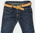 A close-up of dark blue denim jeans with a natural leather belt with a brass Garrison buckle and copper rivets, laid flat against a white background.