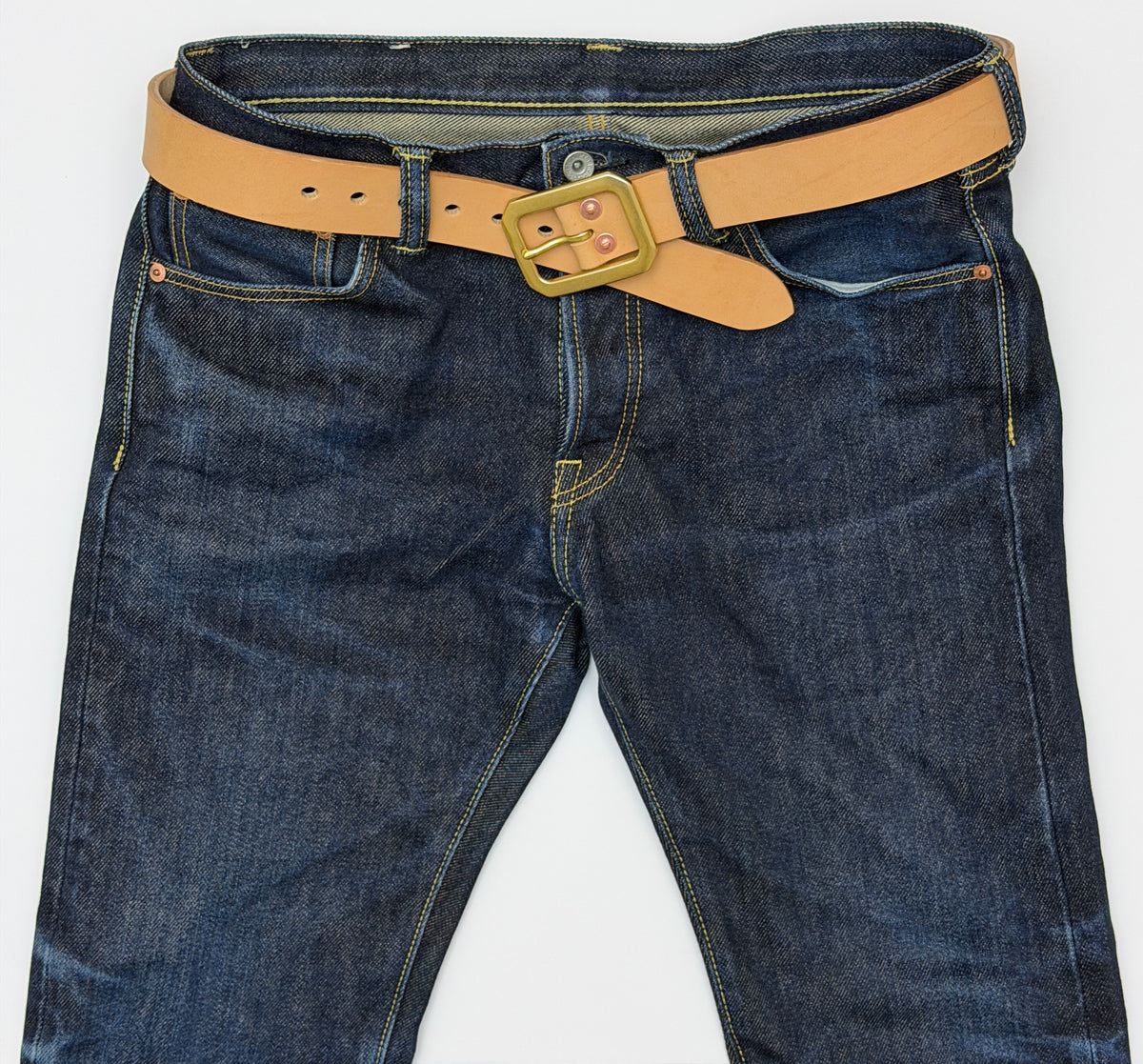 A close-up of dark blue denim jeans with a natural leather belt with a brass Garrison buckle and copper rivets, laid flat against a white background.