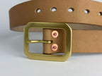 A close-up of a natural leather belt with a large solid brass Garrison buckle and copper rivets, set against a clean white background.