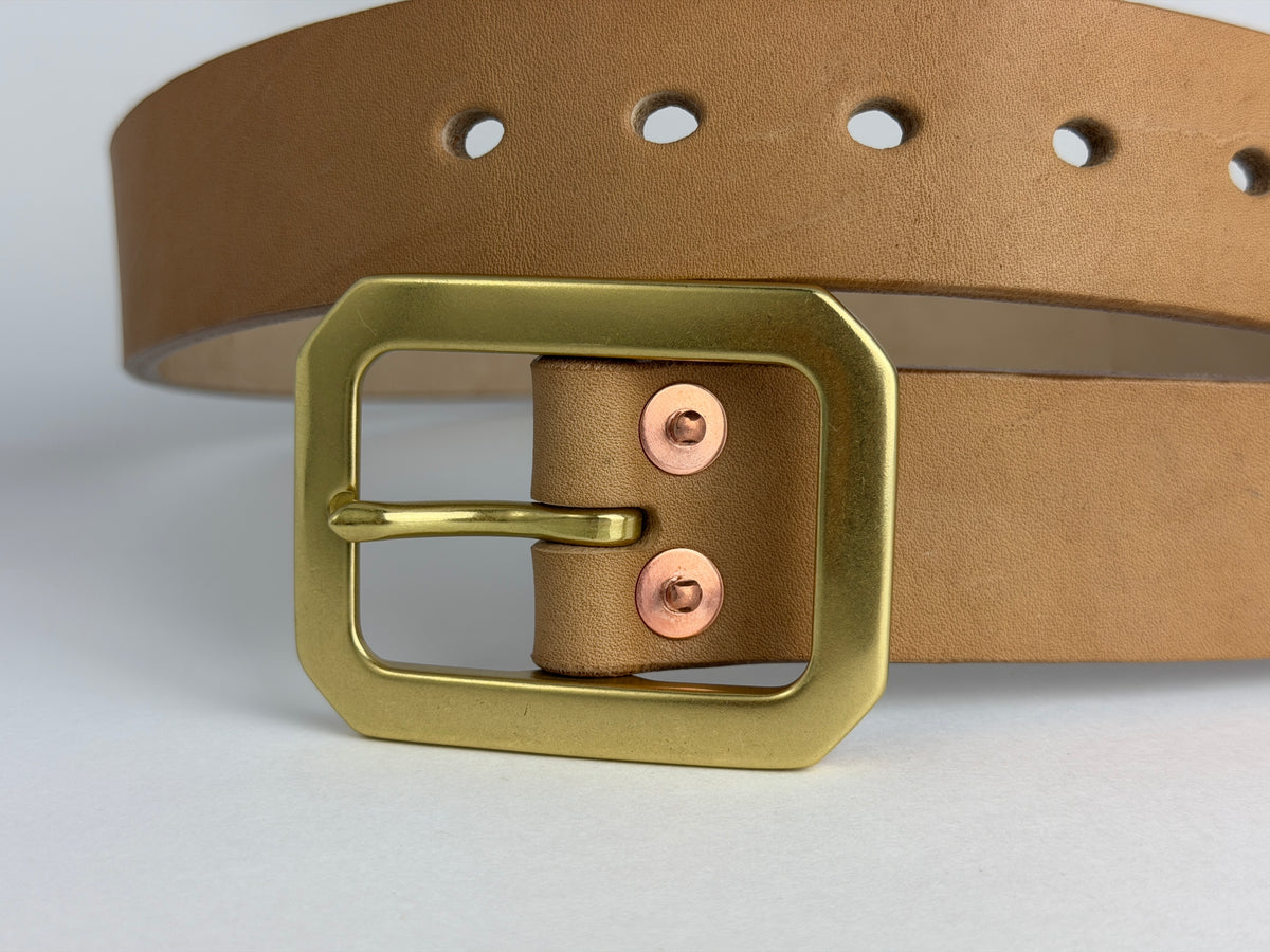 A close-up of a natural leather belt with a large solid brass Garrison buckle and copper rivets, set against a clean white background.