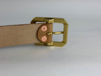 A close-up of the back of a natural brown leather belt with a solid brass metal buckle and red copper rivets, shown on a neutral light gray background.