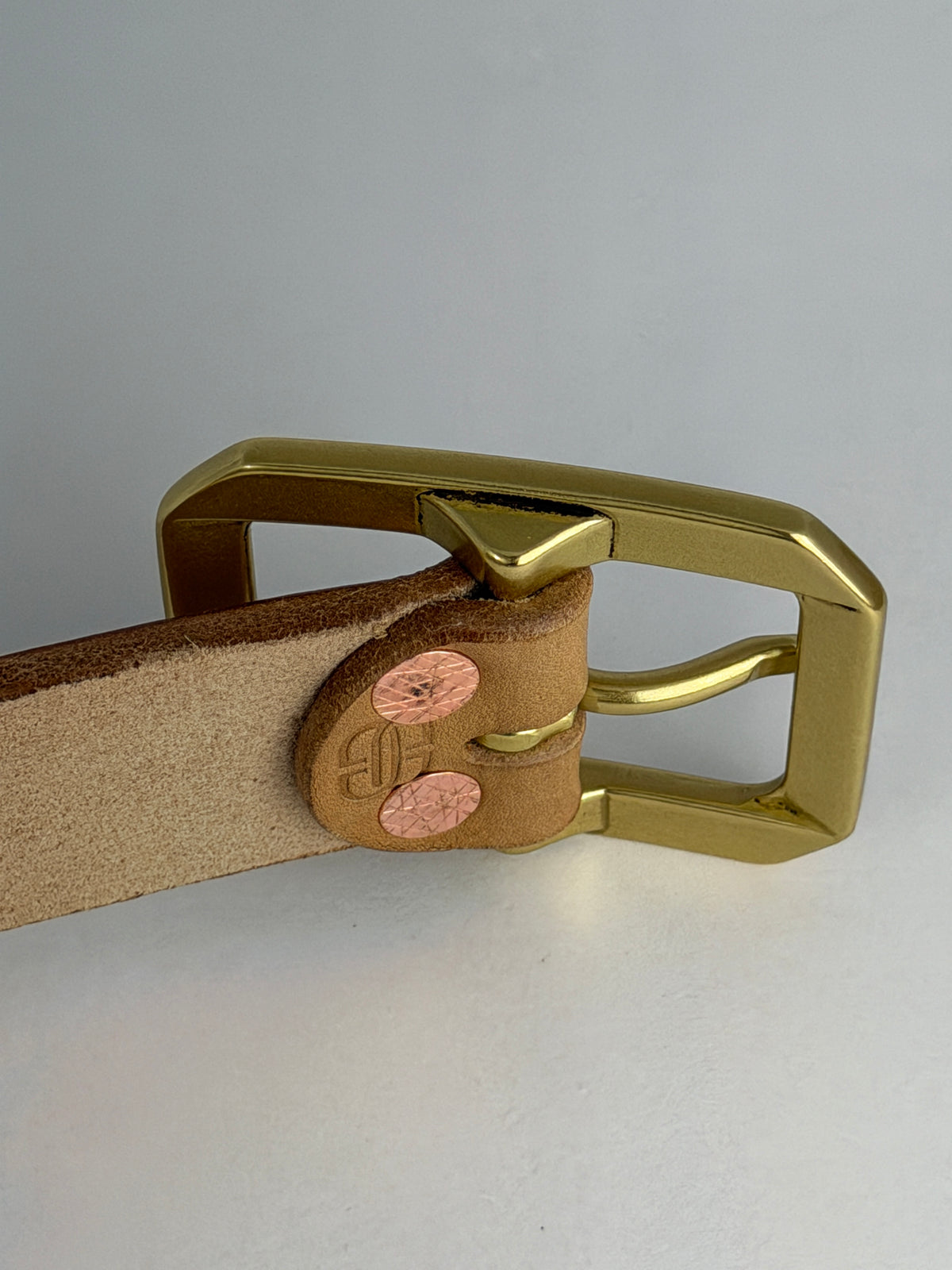 A close-up of the back of a natural brown leather belt, showing a brass buckle and red copper rivets, shown against a light gray background.  There is an embossed stylized "BB" logo on the back.