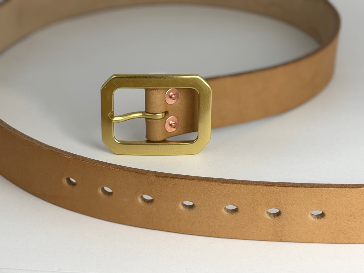 A close-up of a natural brown leather belt with a brass Garrison buckle and red copper rivets, coiled on a clean white background.