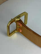 A close-up of a natural leather belt with a brass Garrison buckle and two copper-toned rivets, set against a neutral light gray background.