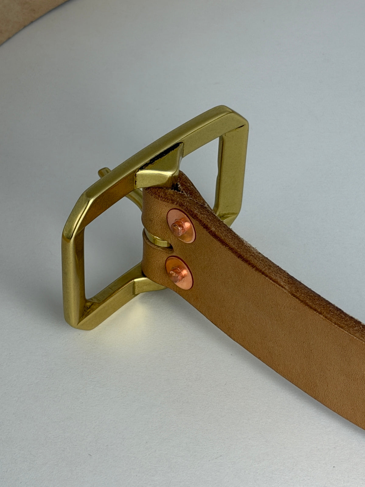 A close-up of a natural leather belt with a brass Garrison buckle and two copper-toned rivets, set against a neutral light gray background.