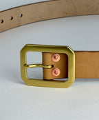 A close-up of a natural leather belt with a large brass Garrison buckle and rose gold-toned rivets, set against a clean light gray background.