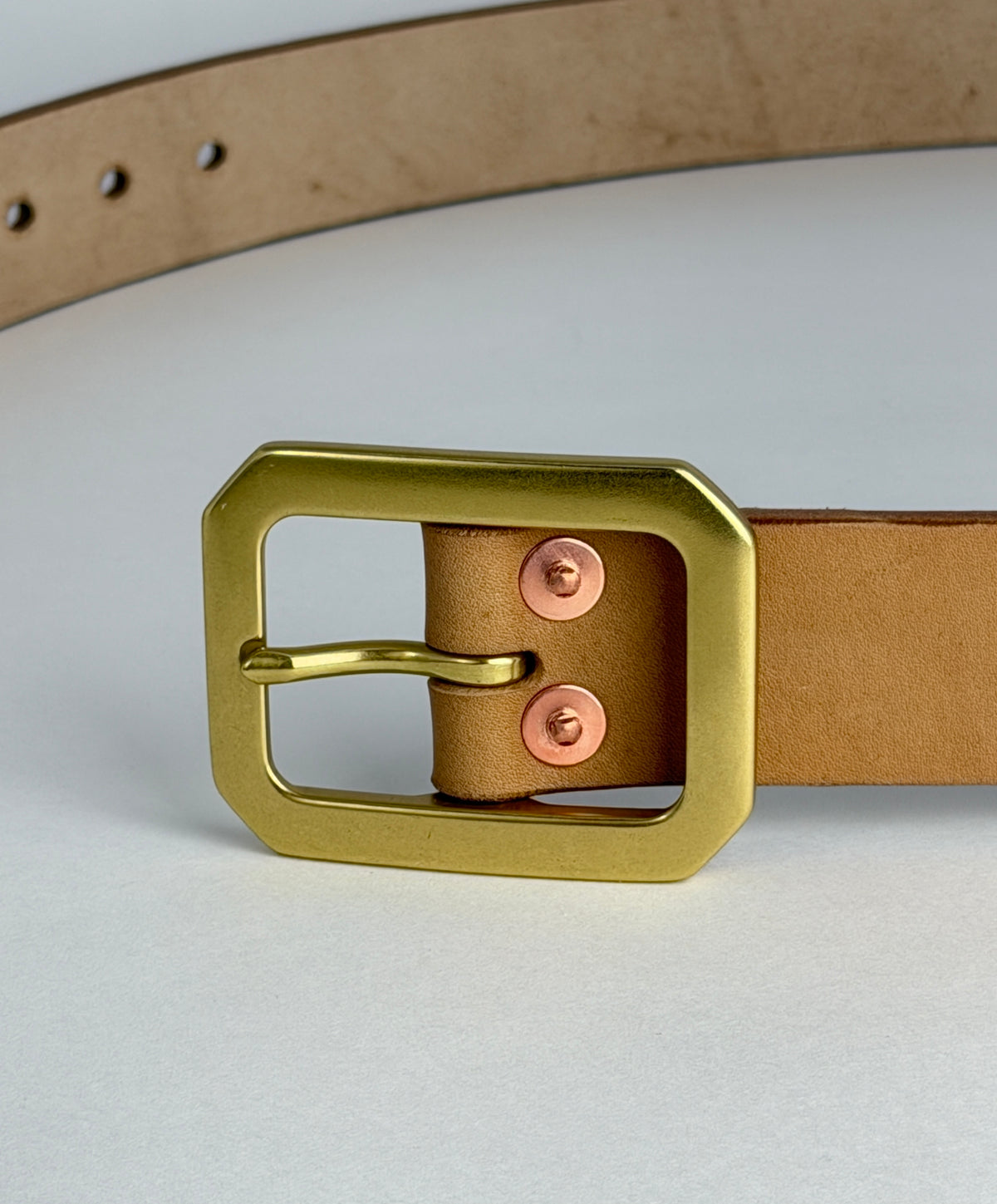 A close-up of a natural leather belt with a large brass Garrison buckle and rose gold-toned rivets, set against a clean light gray background.