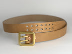 A thick natural leather belt with a large rectangular brass buckle, attached with copper rivets, coiled on a neutral light gray background.