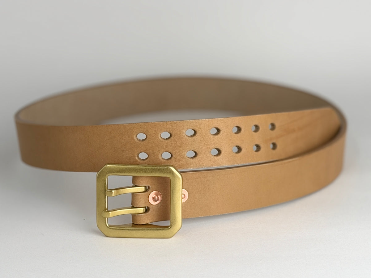 A thick natural leather belt with a large rectangular brass buckle, attached with copper rivets, coiled on a neutral light gray background.