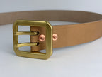 Close up of a natural leather belt with a rectangular dual-pronged brass buckle and rose gold-toned rivets, shown on a light gray background.