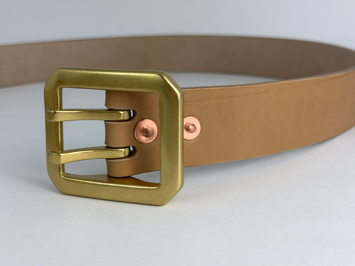 Close up of a natural leather belt with a rectangular dual-pronged brass buckle and rose gold-toned rivets, shown on a light gray background.