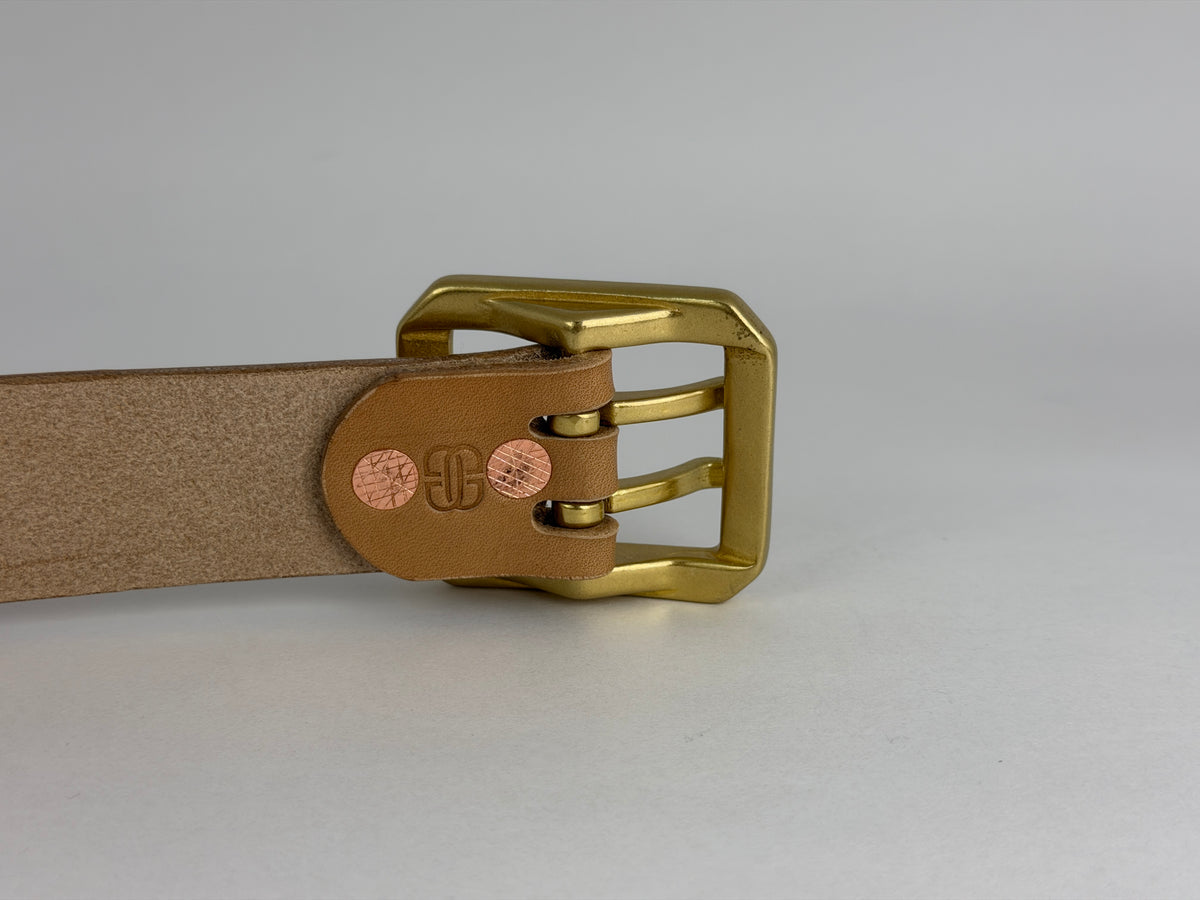 A close-up of the back of a natural leather belt with a polished brass dual-pronged rectangular buckle, showing the inner buckle details and two round copper-colored rivets.