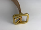 A close-up of a thick natural leather belt with a large rectangular dual-pronged  brass buckle, on a neutral light gray background.