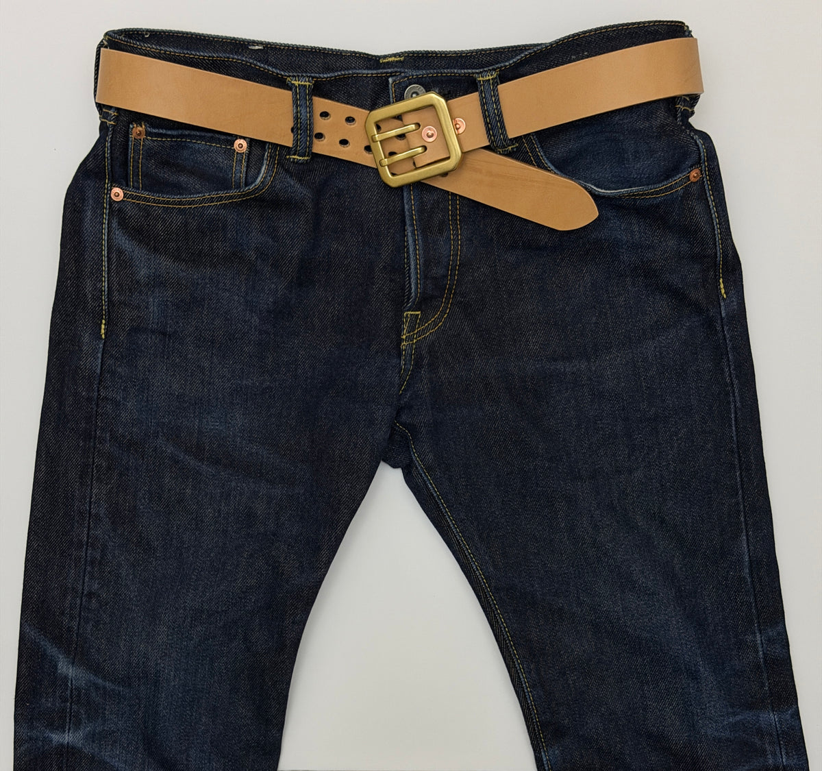 Dark blue denim jeans with a natural leather belt and dual-pronged brass buckle, laid flat against a light gray background.