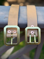 Two natural leather belts resting on a weathered gray wooden surface, with a blurred outdoor background.  The belt on the left has a large rectangular brass buckle with a single prong.  The belt on the right has a more substantial dual-pronged buckle.  Both are attached using copper rivets that are also displayed.