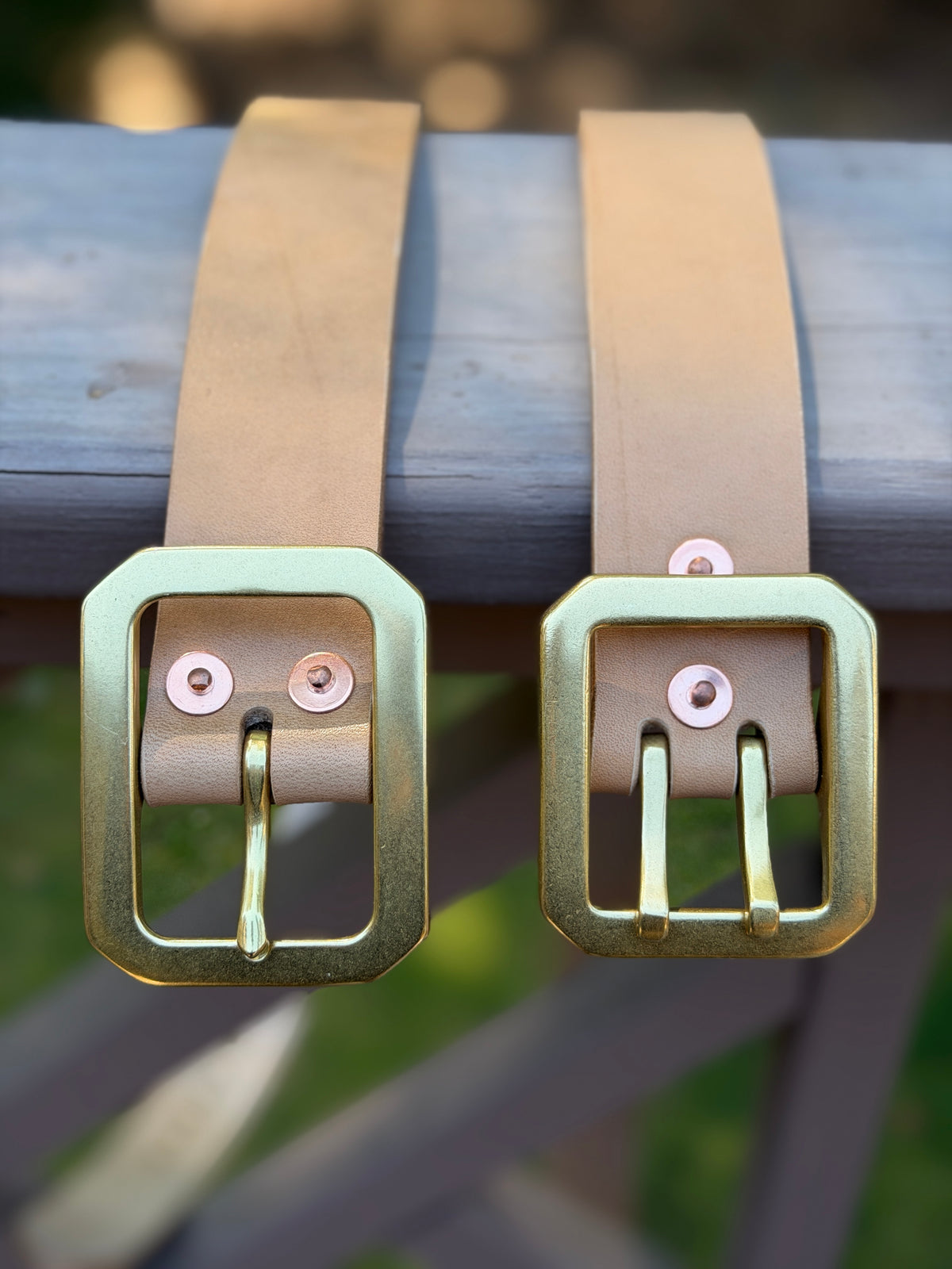 Two natural leather belts resting on a weathered gray wooden surface, with a blurred outdoor background.  The belt on the left has a large rectangular brass buckle with a single prong.  The belt on the right has a more substantial dual-pronged buckle.  Both are attached using copper rivets that are also displayed.