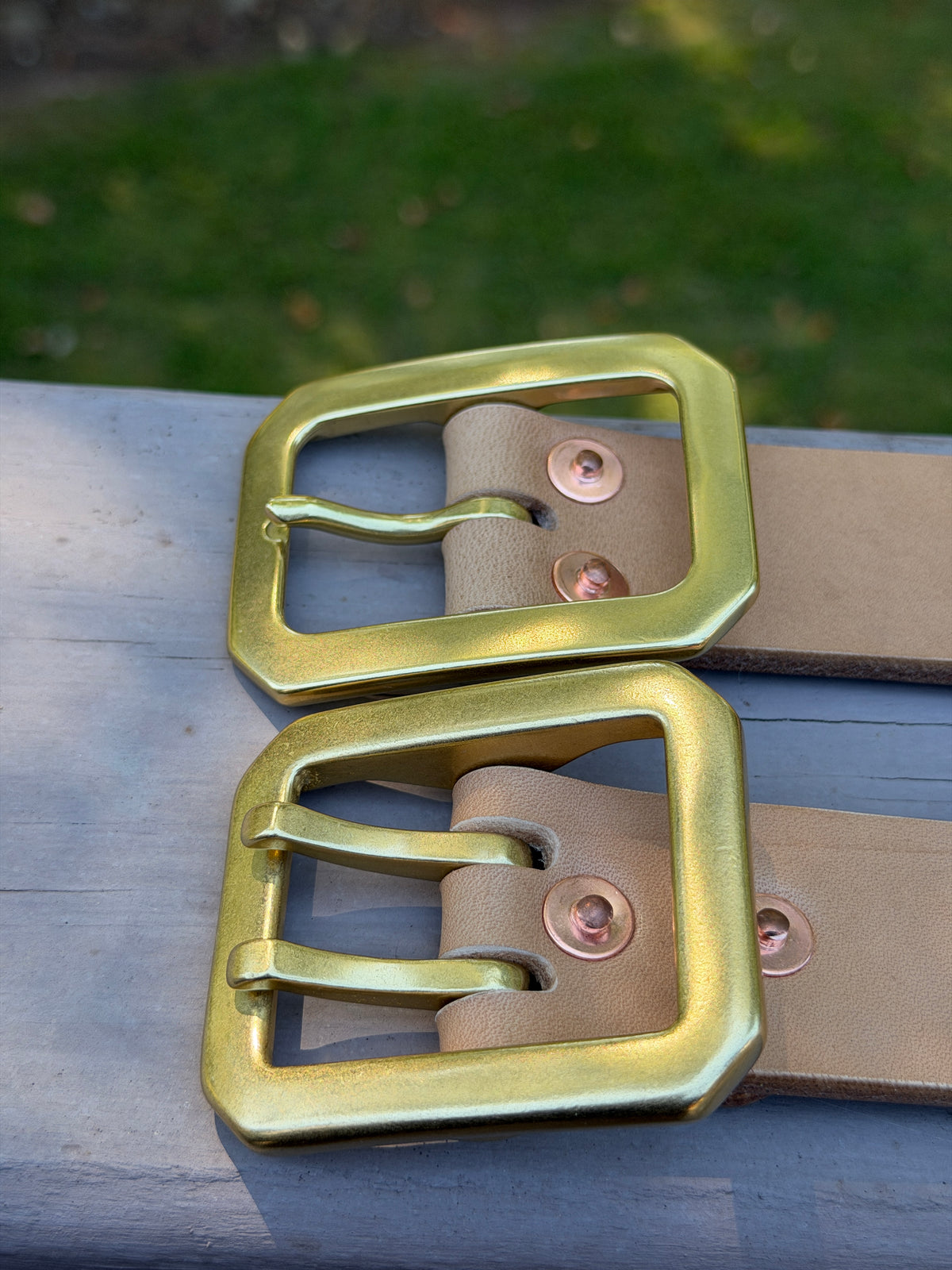 Two natural leather belts resting on a weathered gray wooden surface, with a blurred outdoor background.  The belt on the top has a large rectangular brass buckle with a single prong.  The belt on the bottom has a more substantial dual-pronged buckle.  Both are attached using copper rivets that are also displayed.