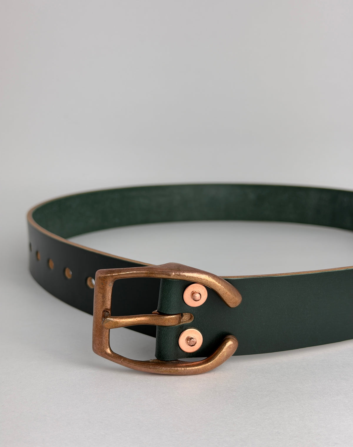 A dark forest green leather belt with a polished copper horseshoe buckle and matching rivets, displayed on a neutral light gray background.