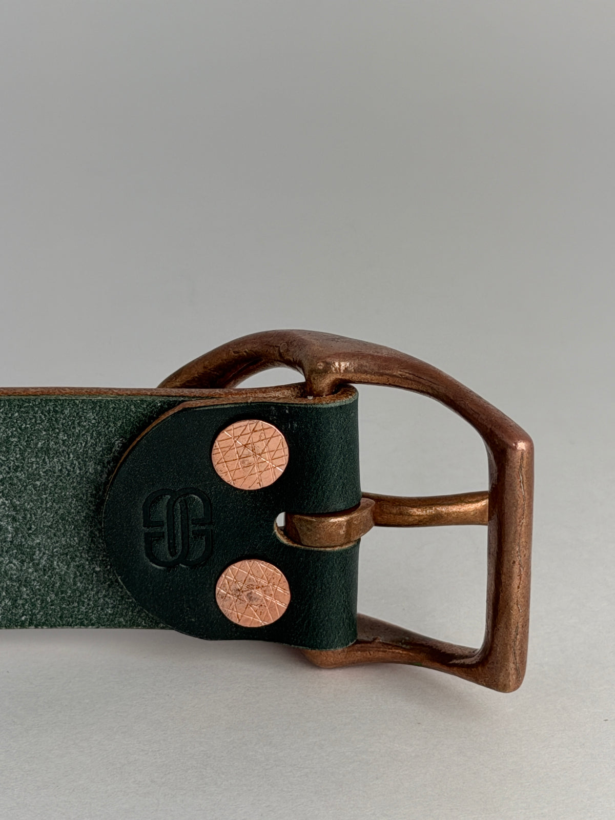 A close-up of the back side of a dark green leather belt, featuring a copper metal horseshoe buckle and two copper rivets, set against a neutral gray background.