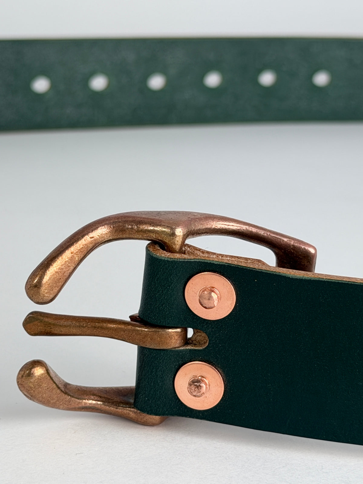 Close-up of a dark green leather belt with a copper horseshoe metal buckle and copper rivets, shown on a light gray background.