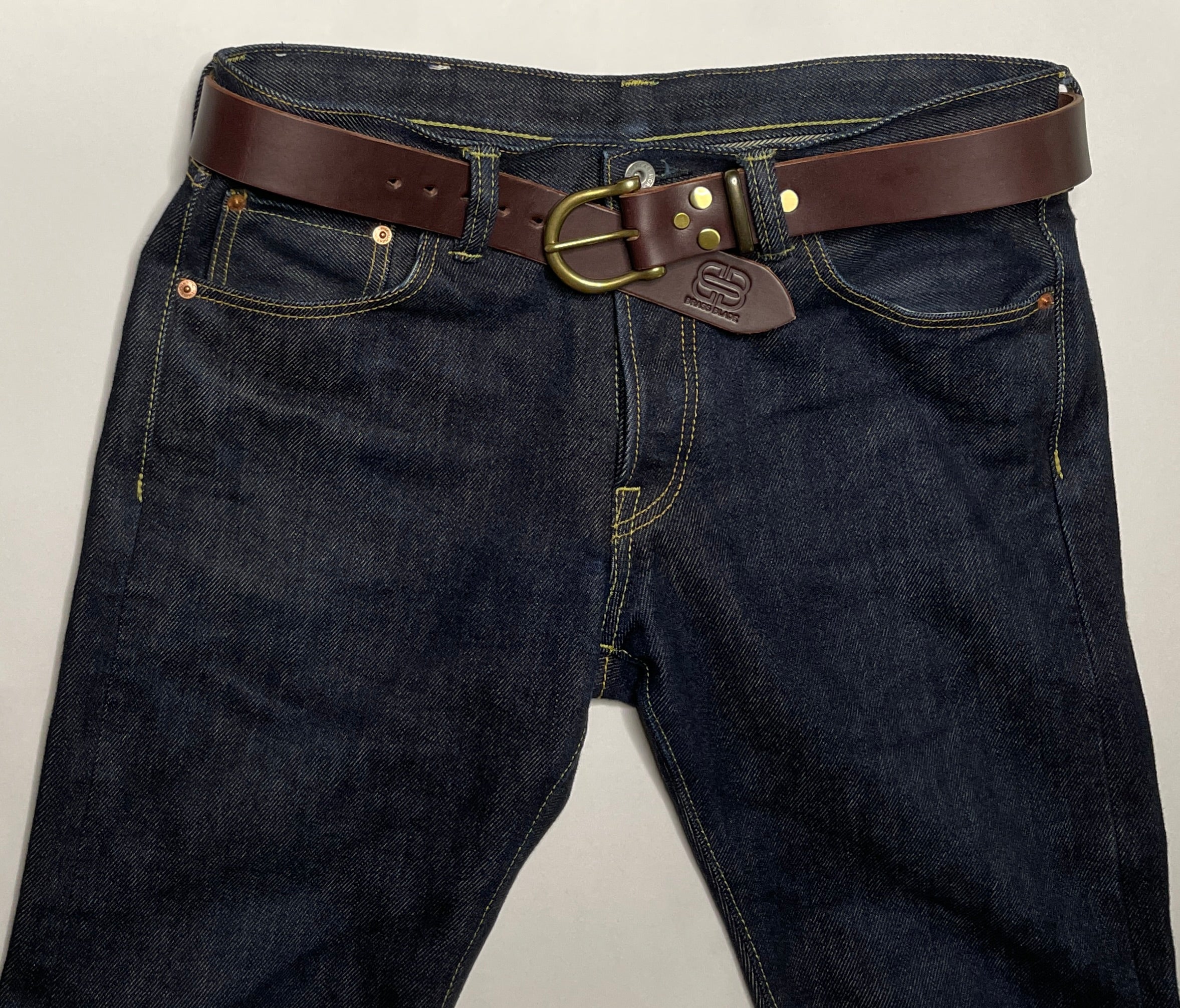 Dark blue jeans with a brown leather belt and brass buckle on a light gray background