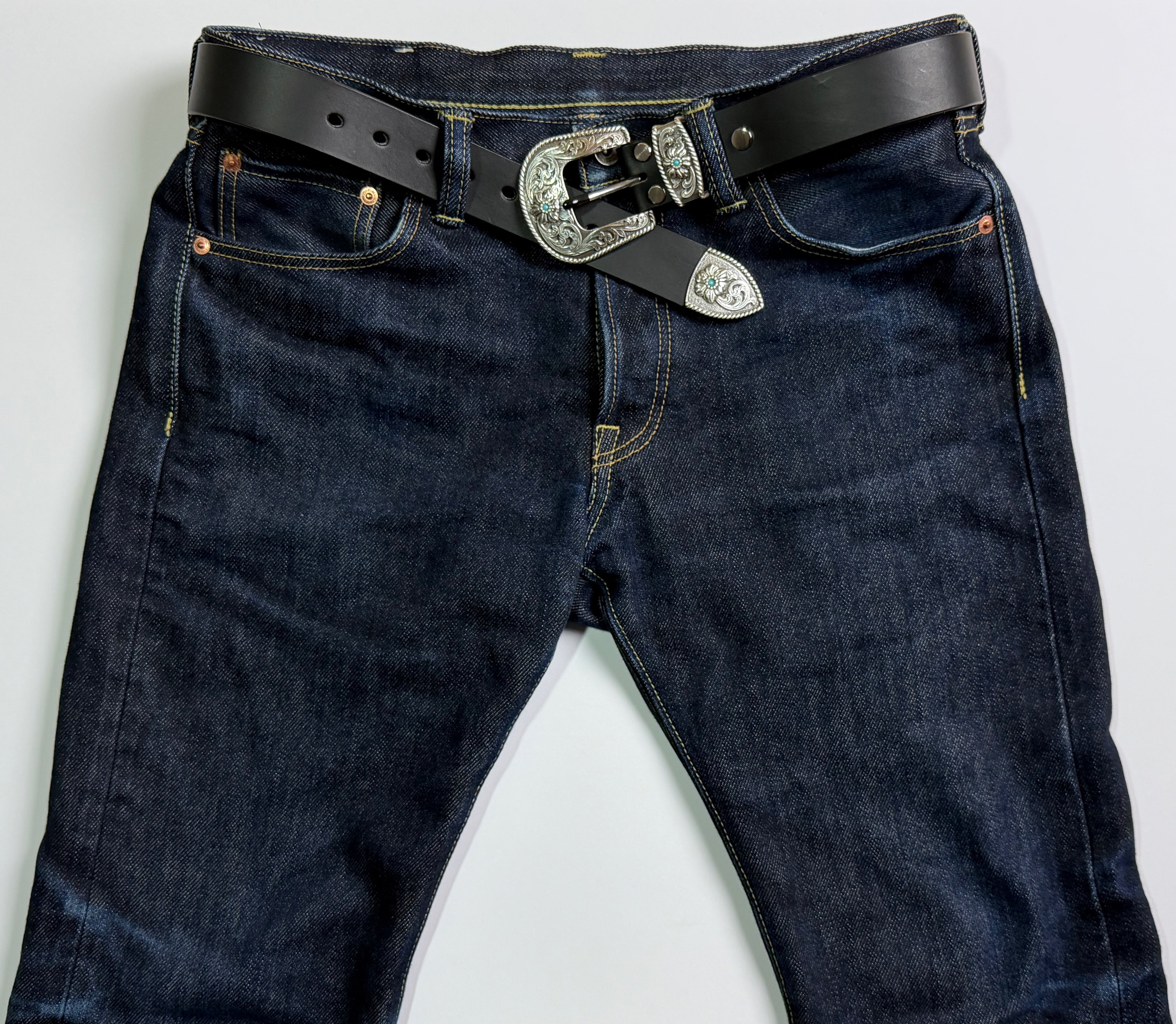 Blue jeans on a white background, with a black western cowboy leather belt with silver buckle and keeper adorned with turquoise stones