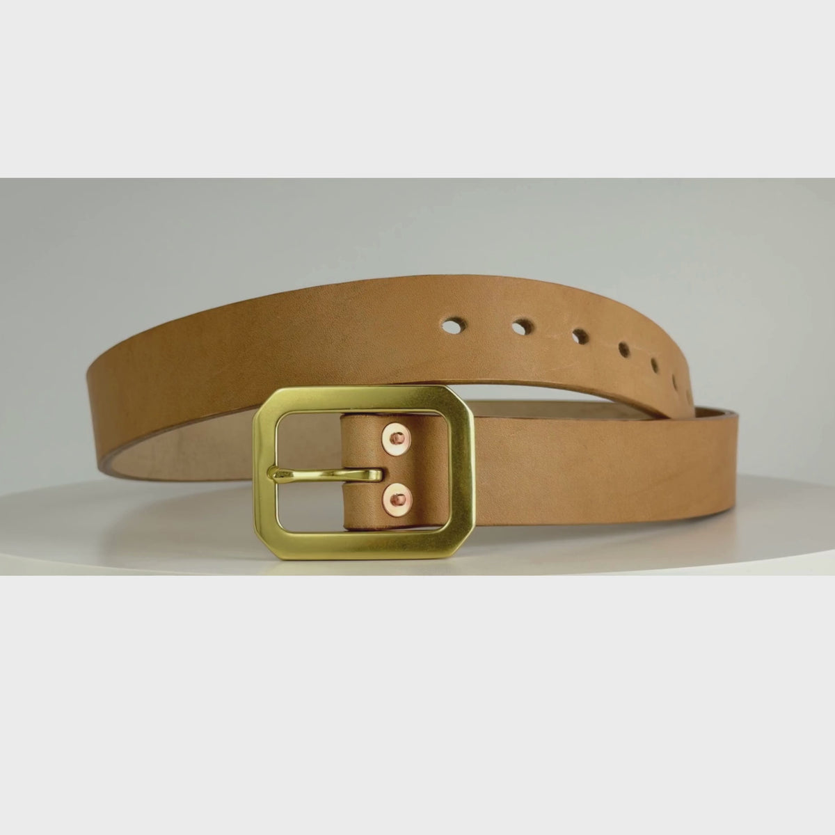 This is a video of a belt rotating on a white carousel.  The belt is made of natural leather, a brass Garrison buckle, and red copper rivets.